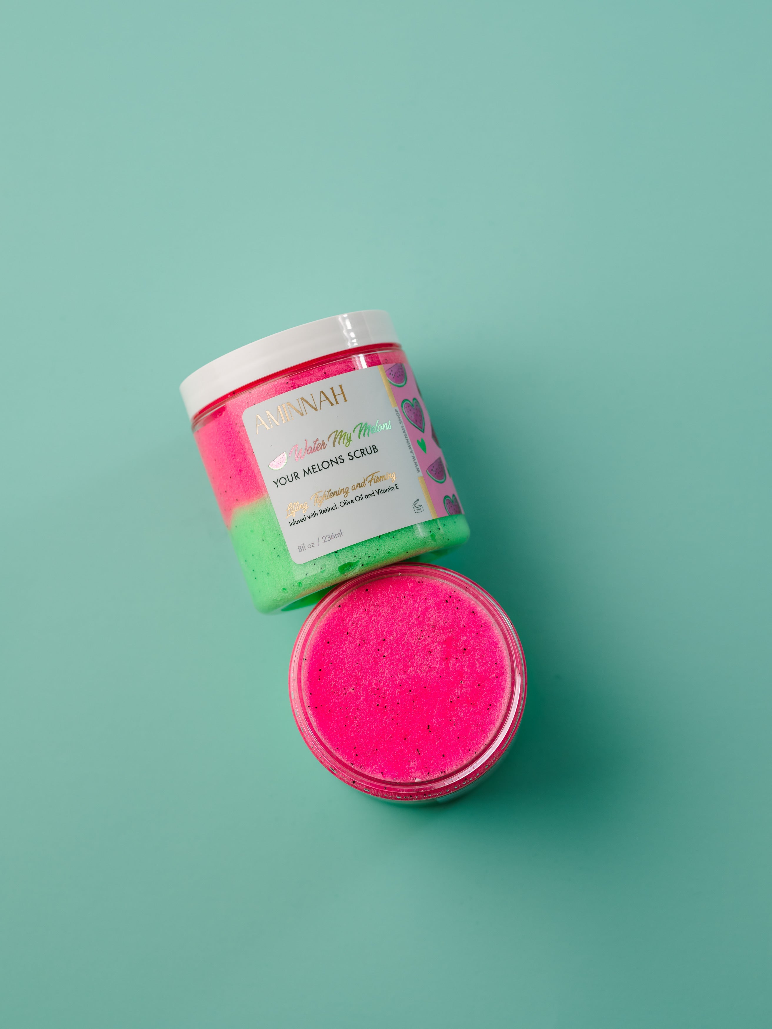 Pink bath scrub in a jar with a visible brand label on a teal background. Water My Melons Boob Sugar Scrub in an open jar with exfoliating pink sugar scrub and branded container on a teal background. Clean beauty skincare made with natural ingredients including watermelon fruit extract, shea butter, cocoa butter, mango butter, aloe vera, vitamin E, retinol, and botanical oils. Cruelty-free, vegan, female-owned beauty brand.