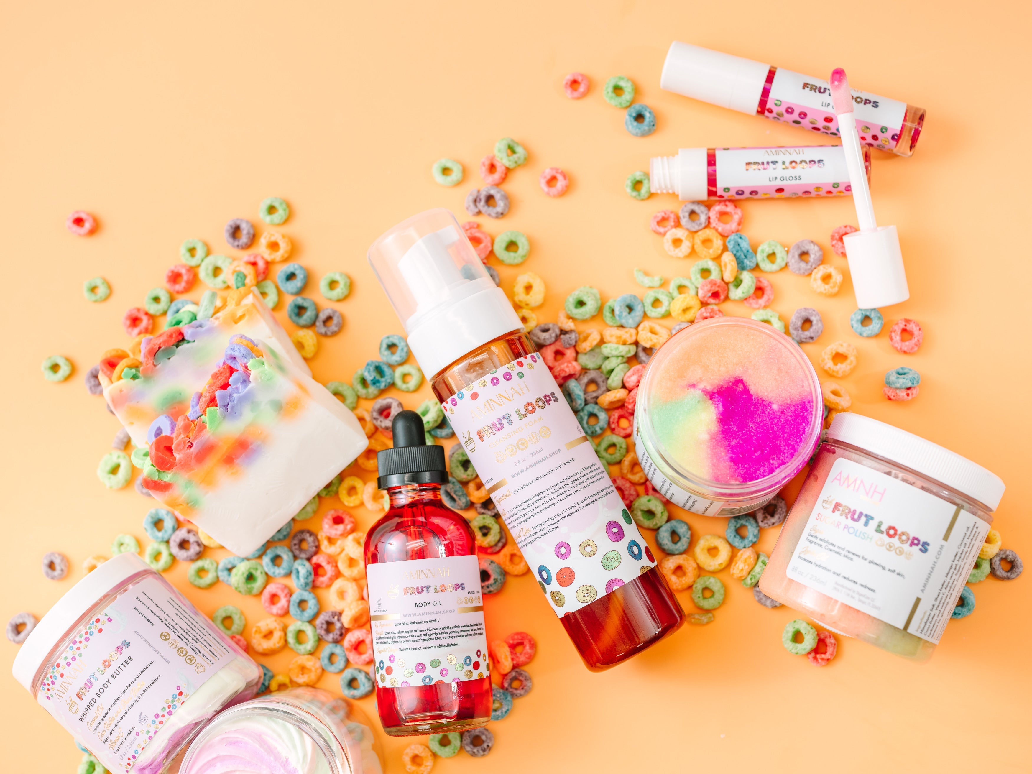 Assorted beauty products including bottles and jars on a colorful background with cereal pieces. AMINNAH "FRUT LOOPS" Body Care Bundle displayed with whipped body butter, sugar scrub, body oil, lip gloss, and handcrafted soap on a bright yellow background surrounded by colorful cereal pieces. Clean beauty, cruelty-free, vegan, femae-owned, skincare collection made with naturally derived ingredients including shea butter, coconut oil, jojoba oil, avocado oil, cocoa butter, rosehip oil, vitamins C and E