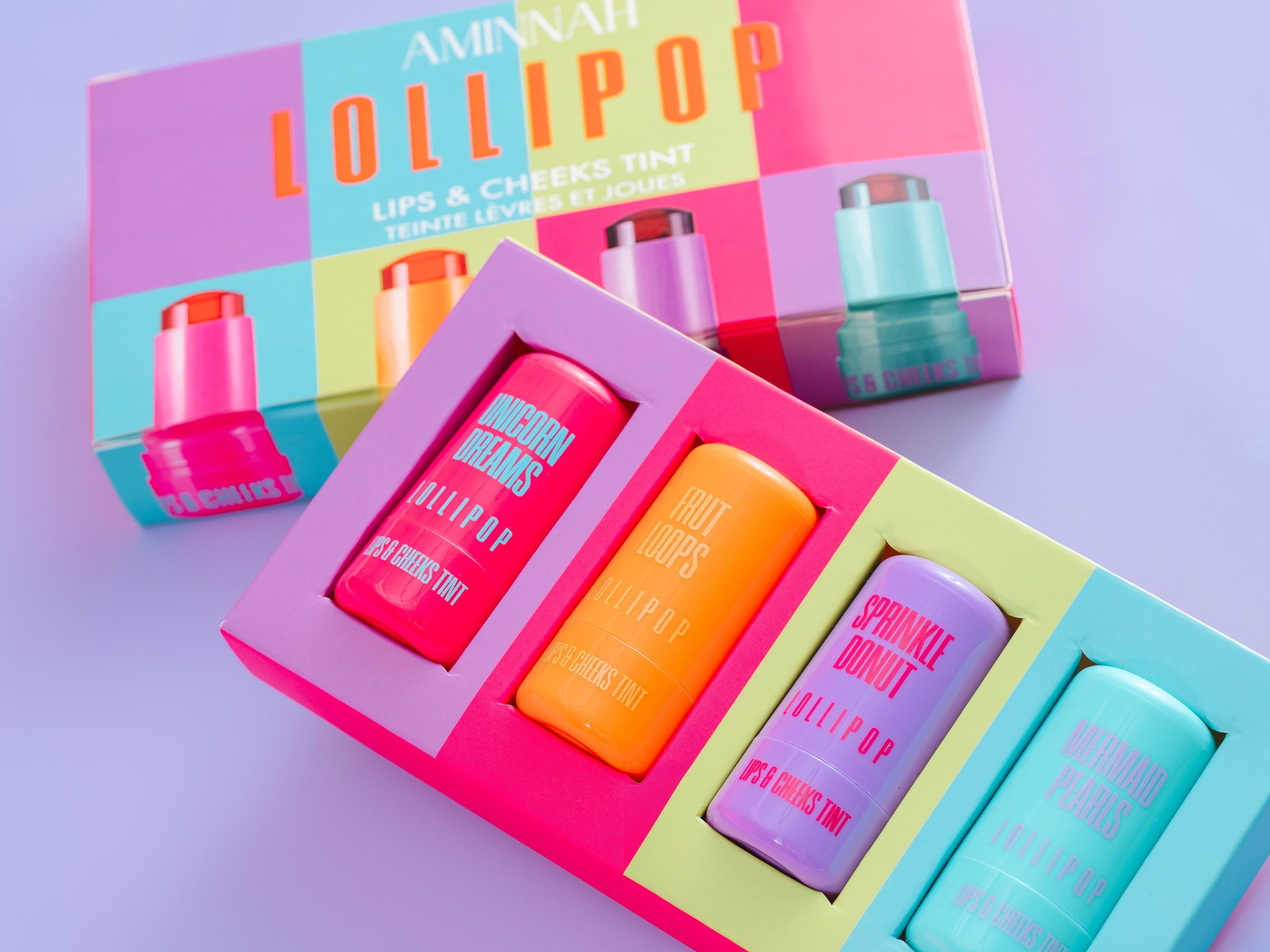 Set of colorful lip products in a box with 'Lollipop' branding on a light purple background. AMINNAH LOLLIPOP Lips & Cheeks Tints Gift Set featuring four colorful hydrating lip and cheek tints made with natural ingredients. Vegan, cruelty-free clean beauty from a female-owned brand in vibrant gift box packaging.
