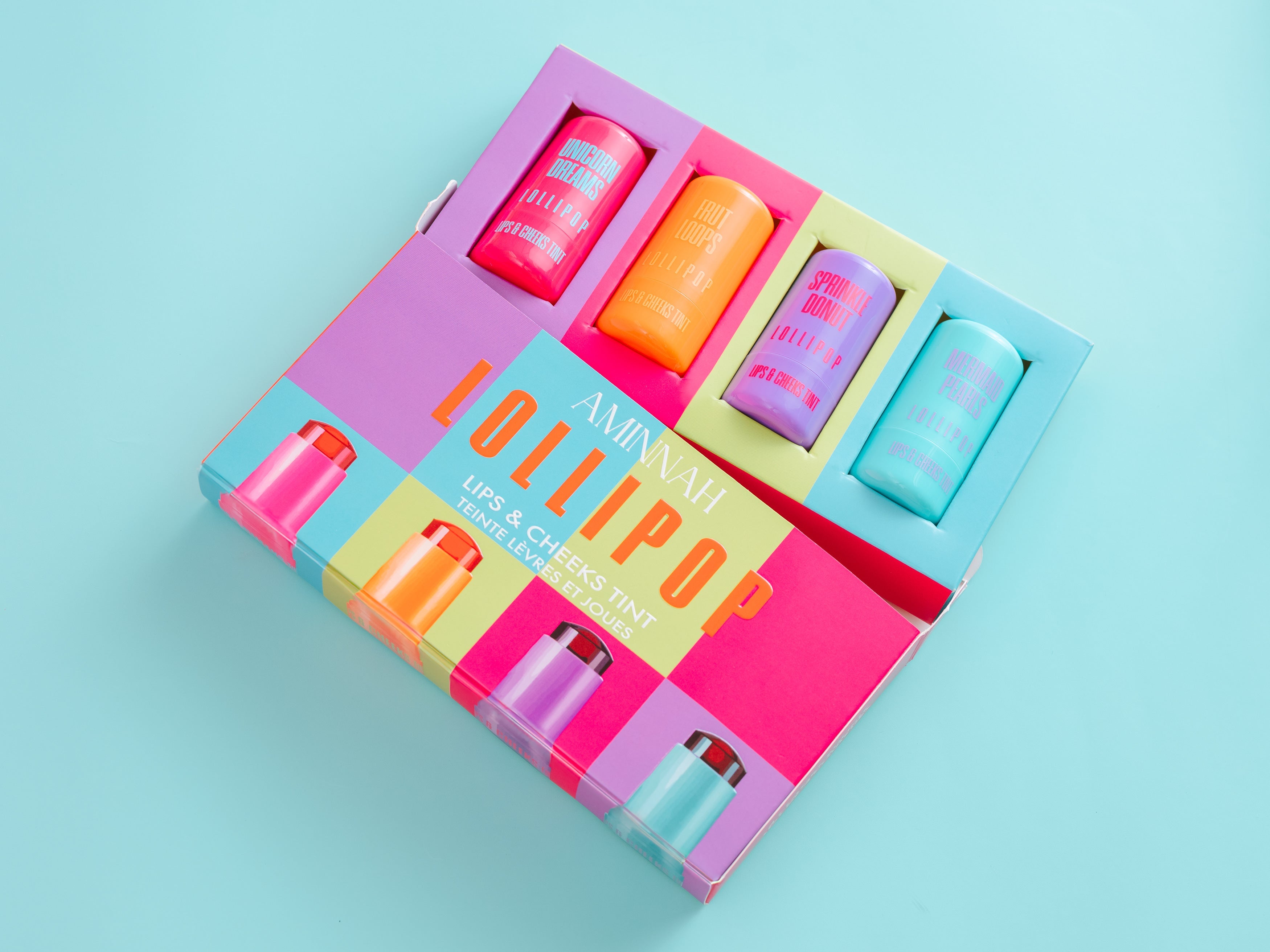 Colorful lip and cheek product set with 'Lollipop' branding on a light blue background. AMINNAH LOLLIPOP Lips & Cheeks Tints Gift Set opened to reveal four colorful lip and cheek tints with vibrant packaging. Vegan, cruelty-free clean beauty made with Aloe Vera, Niacinamide, Hyaluronic Acid, and Goji Berry from a female-owned brand.