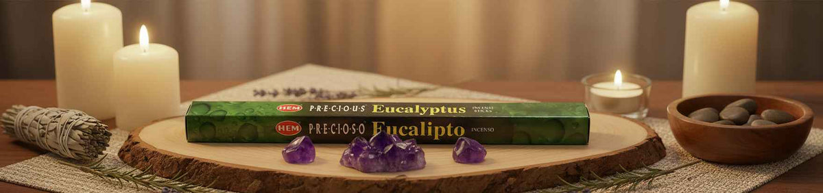 A green box of Preciso Eucalyptus incense sticks is displayed on a wooden slice with purple crystals, surrounded by lit white candles, a sage bundle, and a bowl of stones.