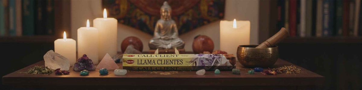A styled tabletop arrangement with lit white candles, crystals, a brass singing bowl, a small Buddha statue, and a book titled 'Llama Clientele' in the foreground, set against a dark, blurred interior background with bookshelves.