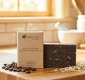 A bar of exfoliating coffee & oatmeal soap stands in front of its kraft paper packaging box on a wooden cutting board, with coffee beans and oat flakes scattered around it. The background shows a softly blurred kitchen scene with warm light coming through a window.
