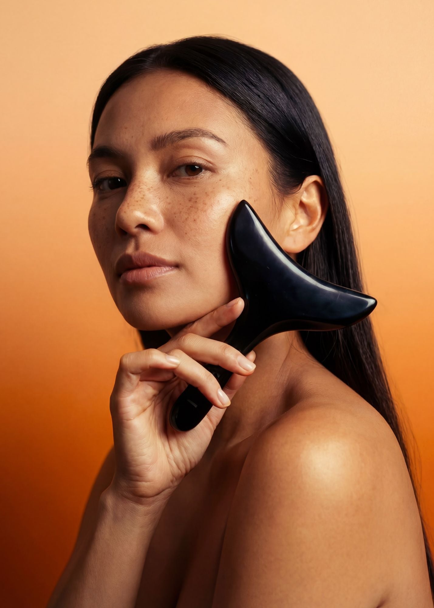 Woman using a black facial tool on her face against an orange background. The Lymphatic Detox System