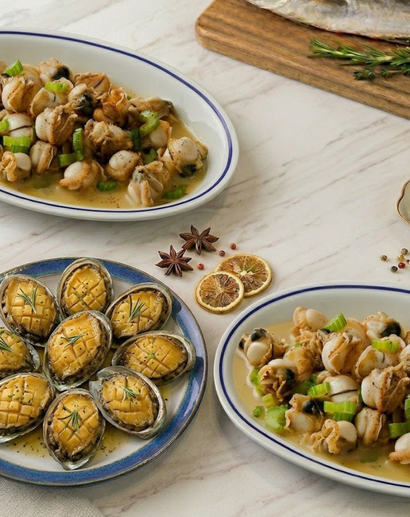 Three plates of prepared seafood scallops on a light marble-style surface: a plate of abalone shells, a plate of mixed shellfish with green onions, and another plate of shellfish in sauce. Spices and dried citrus slices are arranged around them, with a wooden board in the background.