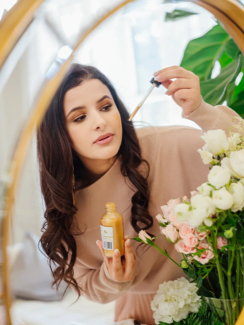 24K Gold Glow Body Oil reflected in a vanity mirror as a woman applies the nourishing oil with a dropper. Luxurious body oil made with natural ingredients to hydrate, soften, and illuminate skin with a radiant glow. Clean beauty, cruelty-free, vegan skincare from a female-owned brand for healthy, glowing skin.