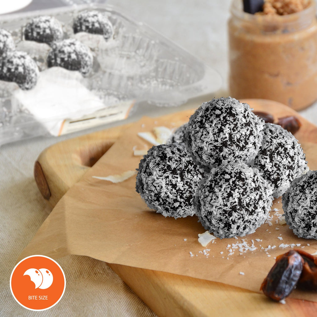 Coconut-coated sugar-free chocolate rum balls on a wooden serving board with dates and a blurred jar in the background.
