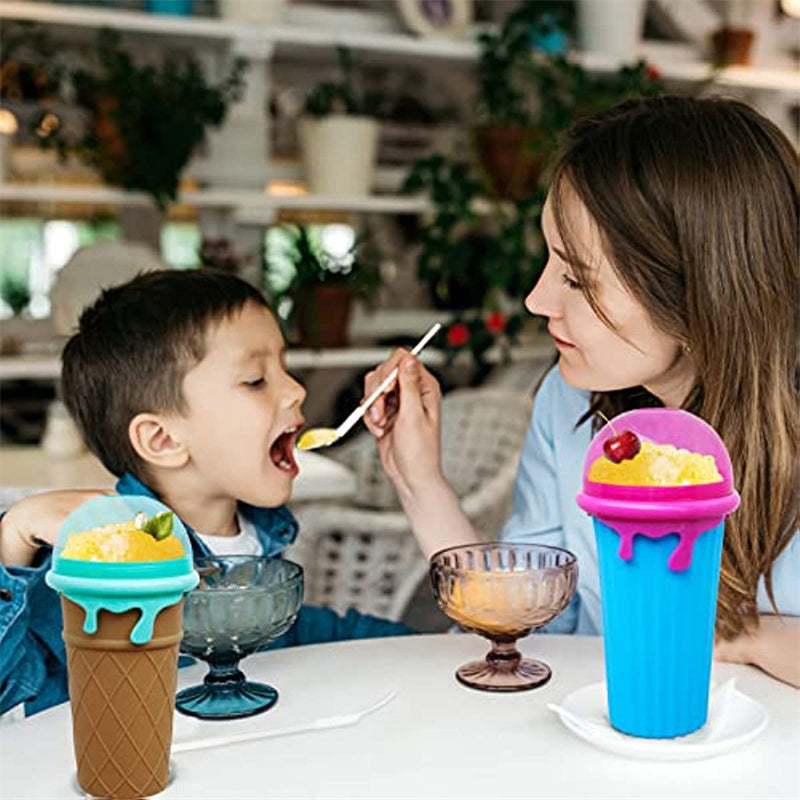 Mother feeding a young child a homemade frozen slush from a reusable ice cream-shaped slushy maker cup with a spoon at a kitchen table, showcasing a fun, family-friendly smoothie maker.