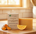 Aspen Kay Naturals handcrafted soap bar in a clear container next to its kraft paper box, styled on a wooden cutting board with fresh turmeric roots and an orange slice in a bright kitchen setting.
