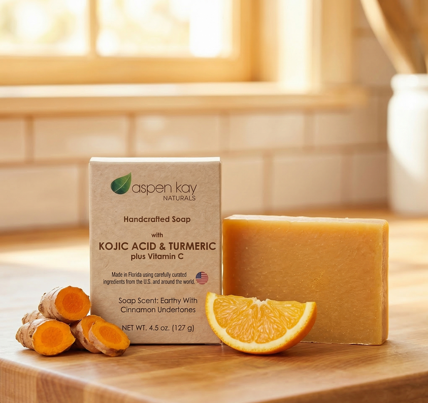 Aspen Kay Naturals handcrafted soap bar in a clear container next to its kraft paper box, styled on a wooden cutting board with fresh turmeric roots and an orange slice in a bright kitchen setting.