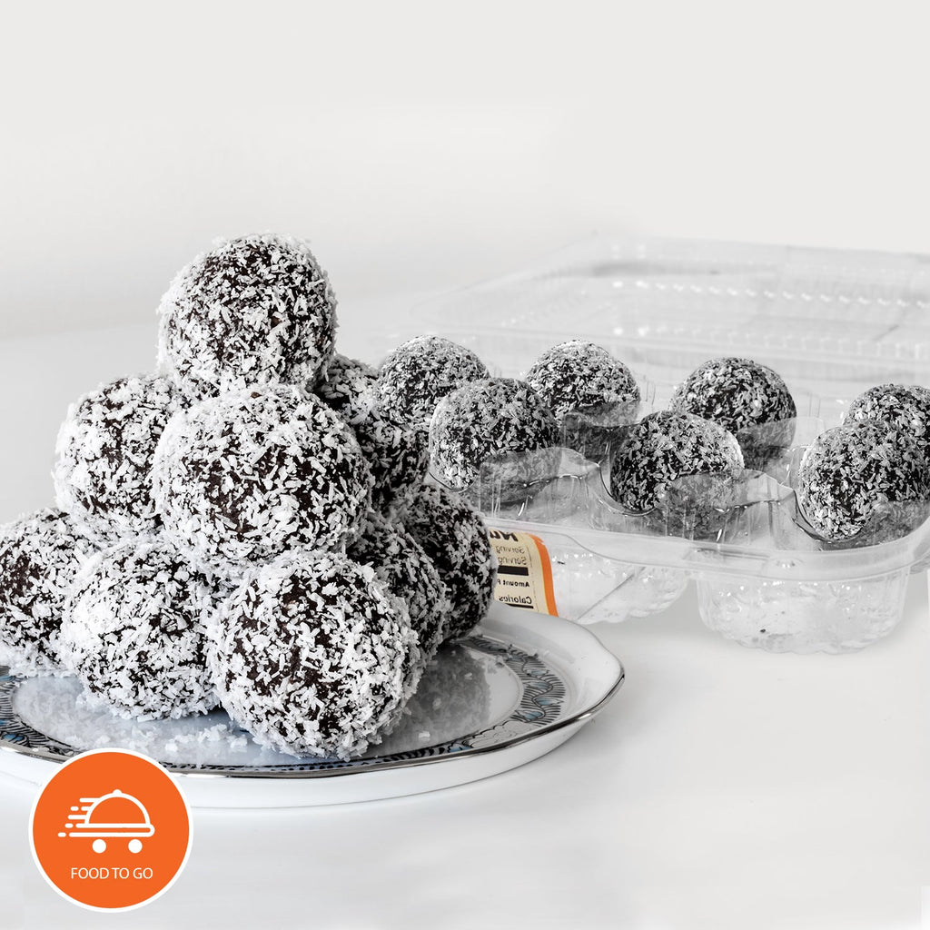 Chocolate rum balls coated in shredded coconut on a decorative plate with additional keto-friendly rum balls in a clear plastic tray on a white background.