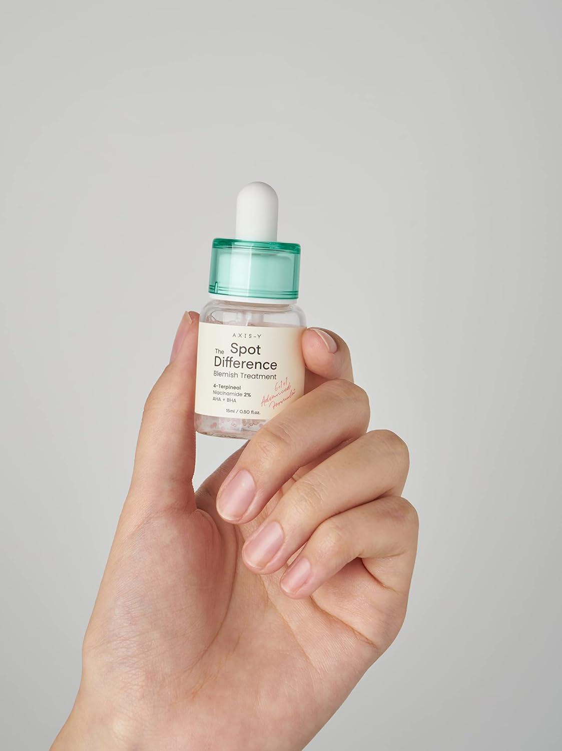 Hand holding the AXIS-Y Spot The Difference Blemish Treatment serum bottle with a green dropper cap against a clean gray background.