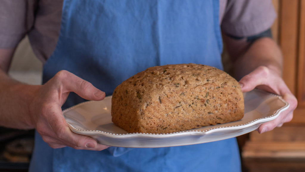 Freshly baked gluten-free rosemary and olive oil sourdough loaf presented on a white serving platter, held by a person wearing a blue apron in a rustic kitchen.