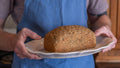 Freshly baked gluten-free rosemary and olive oil sourdough loaf presented on a white serving platter, held by a person wearing a blue apron in a rustic kitchen.