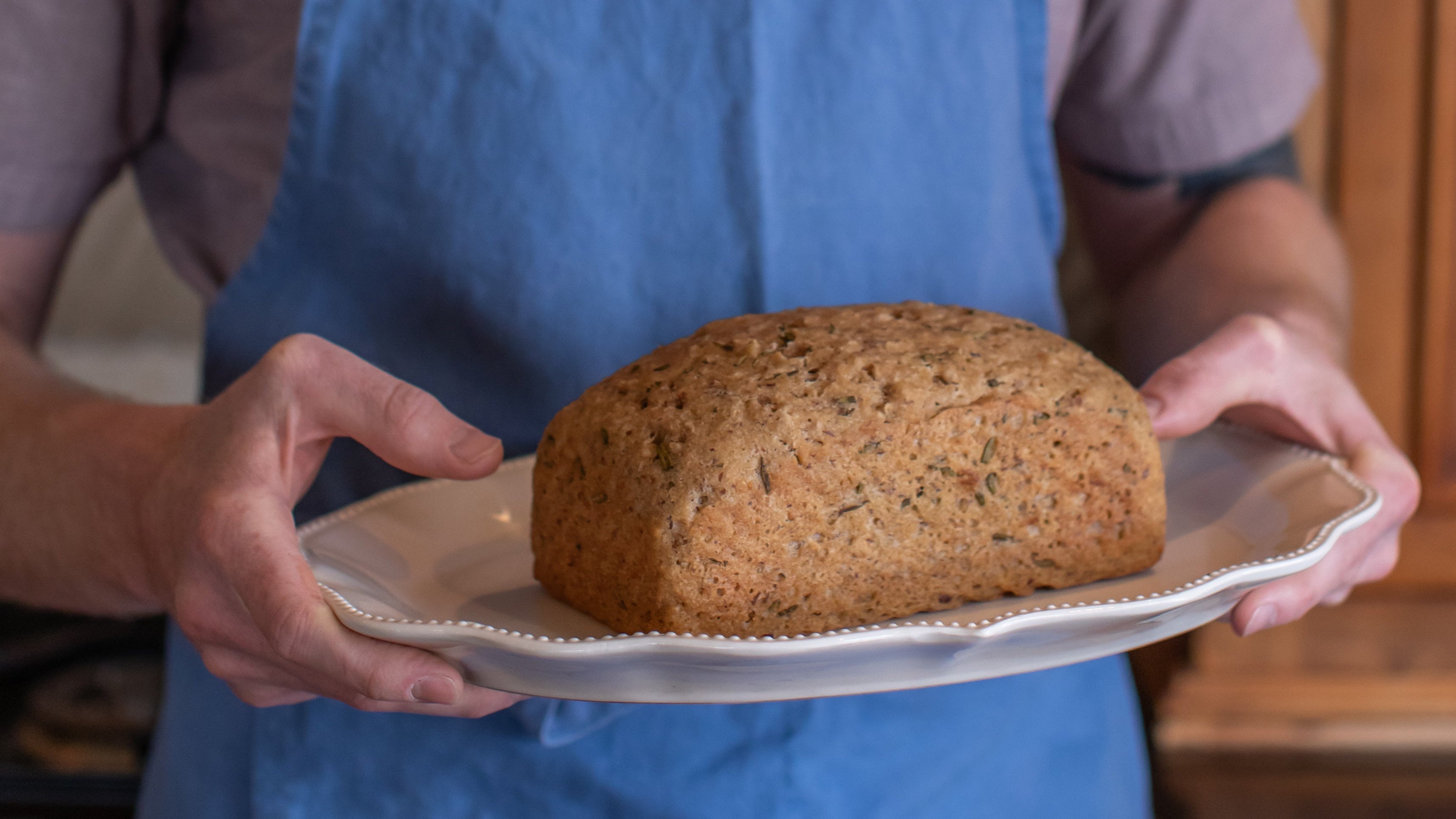 Freshly baked gluten-free rosemary and olive oil sourdough loaf presented on a white serving platter, held by a person wearing a blue apron in a rustic kitchen.