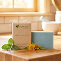 Aspen Kay Naturals Arnica & Mint soap bar in a light blue speckled finish, displayed next to its kraft paper packaging box on a wooden surface with mint leaves and yellow flowers.