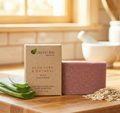 A bar of Aspen Kay Naturals Aloe Vera & Oatmeal with Calamine soap stands next to its kraft paper packaging box on a wooden surface, with aloe vera leaves and rolled oats as props in a warm, softly blurred kitchen setting.