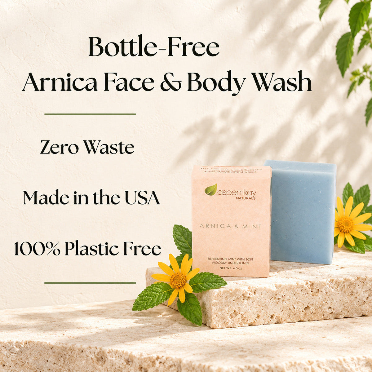 Aspen Kay Naturals Arnica & Mint face and body wash shown as a pale pink box beside a light blue rectangular bar soap, styled on a stone surface with yellow flowers and green leaves against a sunlit beige wall.