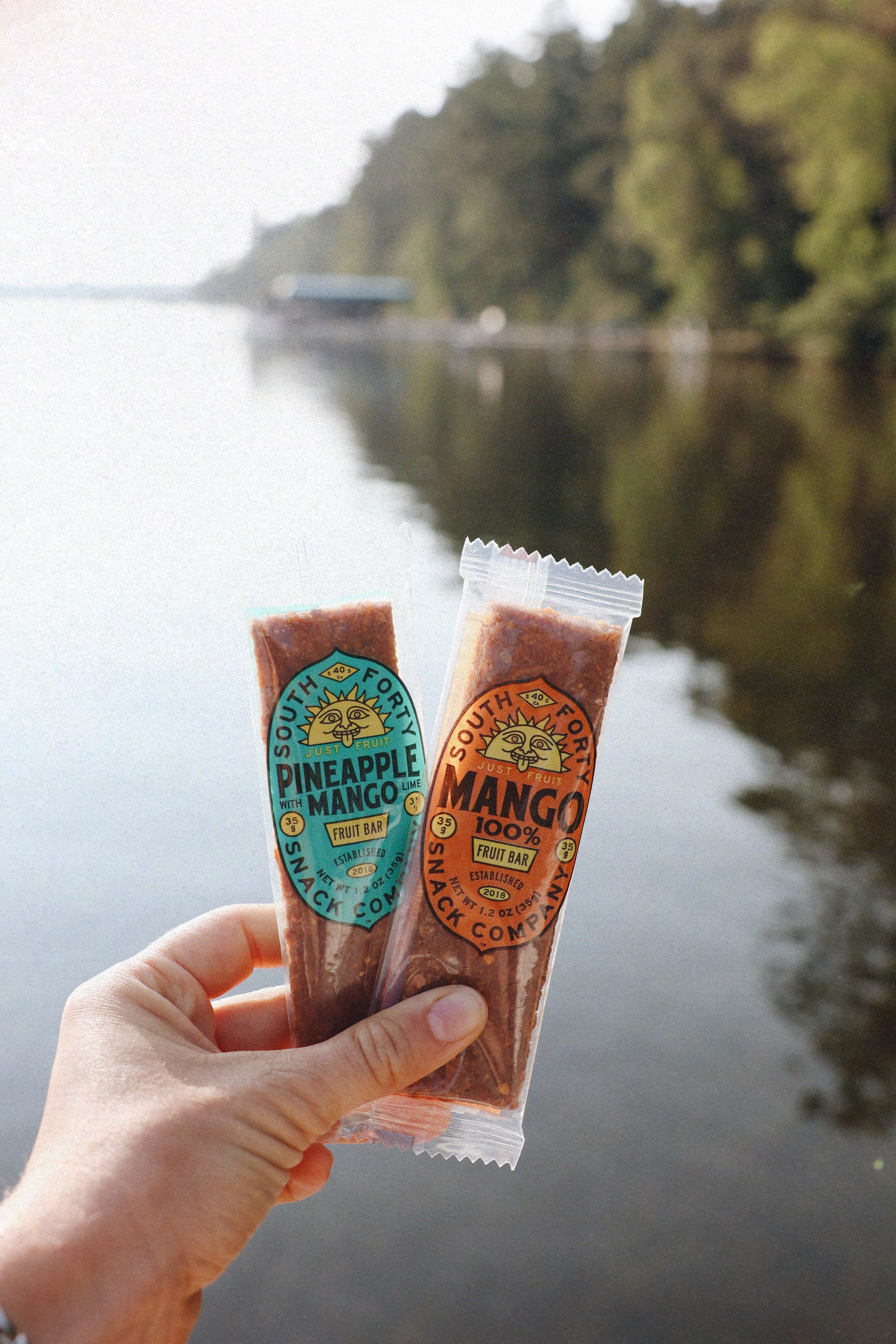 South Forty Pineapple Mango and 100% Mango fruit bars held in hand by a peaceful lake, all-natural tropical dried fruit snacks, vegan, gluten-free, one-ingredient healthy snack bars, portable on-the-go fruit snacks, Product of Ghana and Ivory Coast, outdoor adventure snack, wholesome non-GMO fruit bars, clean ingredient tropical fruit treats.