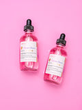 Two bottles of pink liquid with black droppers on a pink background. Two bottles of AMINNAH Unicorn Dreams Body Oil with dropper applicators displayed on a pink background. Clean beauty, cruelty-free, vegan body oil made with natural ingredients including Golden Jojoba Oil, Coconut Oil, Rosehip Oil, Avocado Oil, Sunflower Oil, Hazelnut Oil, and Vitamin E from a female-owned brand.