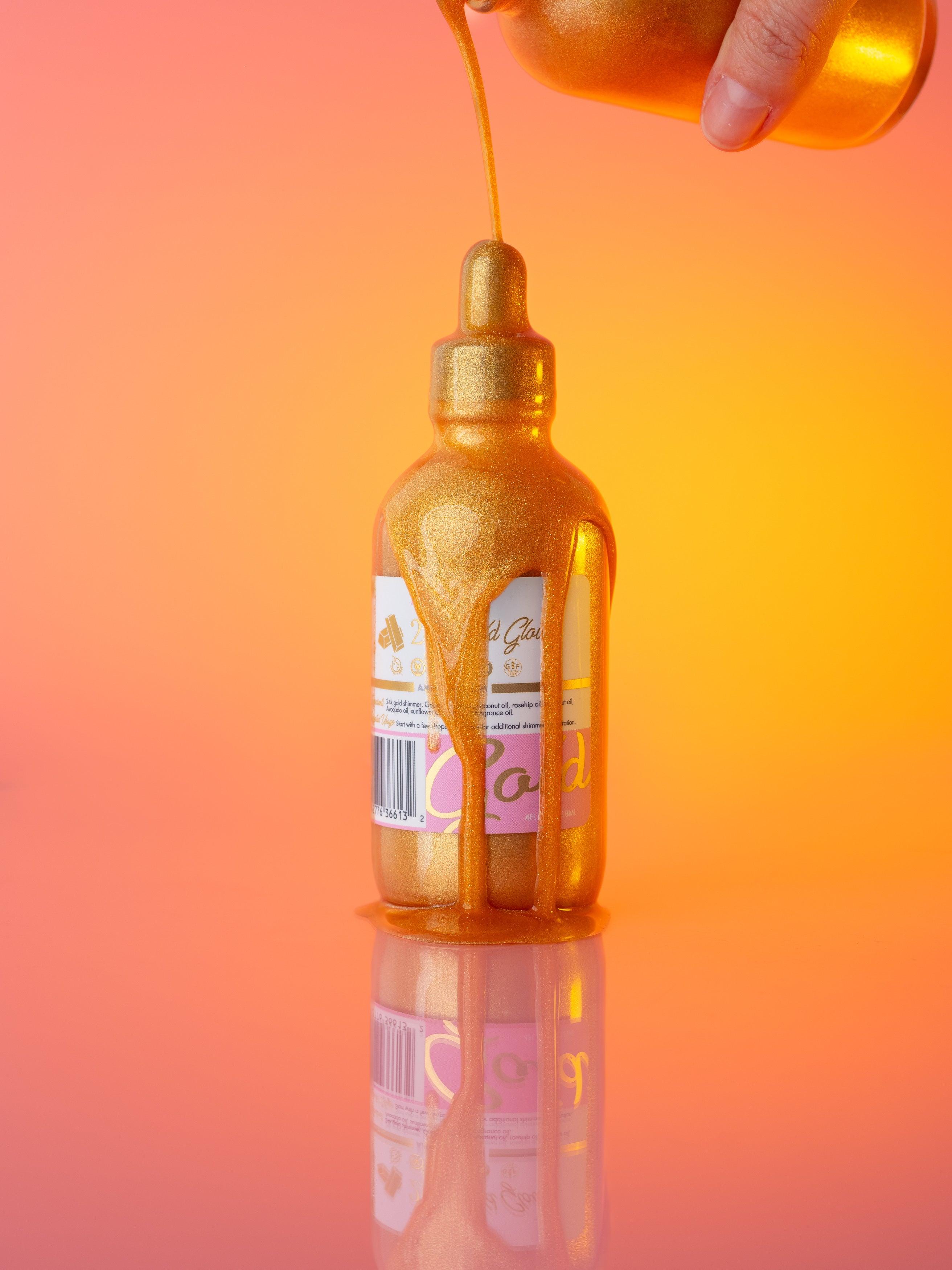 AMINNAH 24K Gold Glow Body Oil bottle with shimmering golden body oil being poured from the dropper against a warm peach and gold background. Clean beauty, cruelty-free, vegan body oil made with natural ingredients including Golden Jojoba Oil, Coconut Oil, Rosehip Oil, Avocado Oil, Sunflower Oil, Vitamin E, and 24K Gold shimmer from a female-owned brand.
