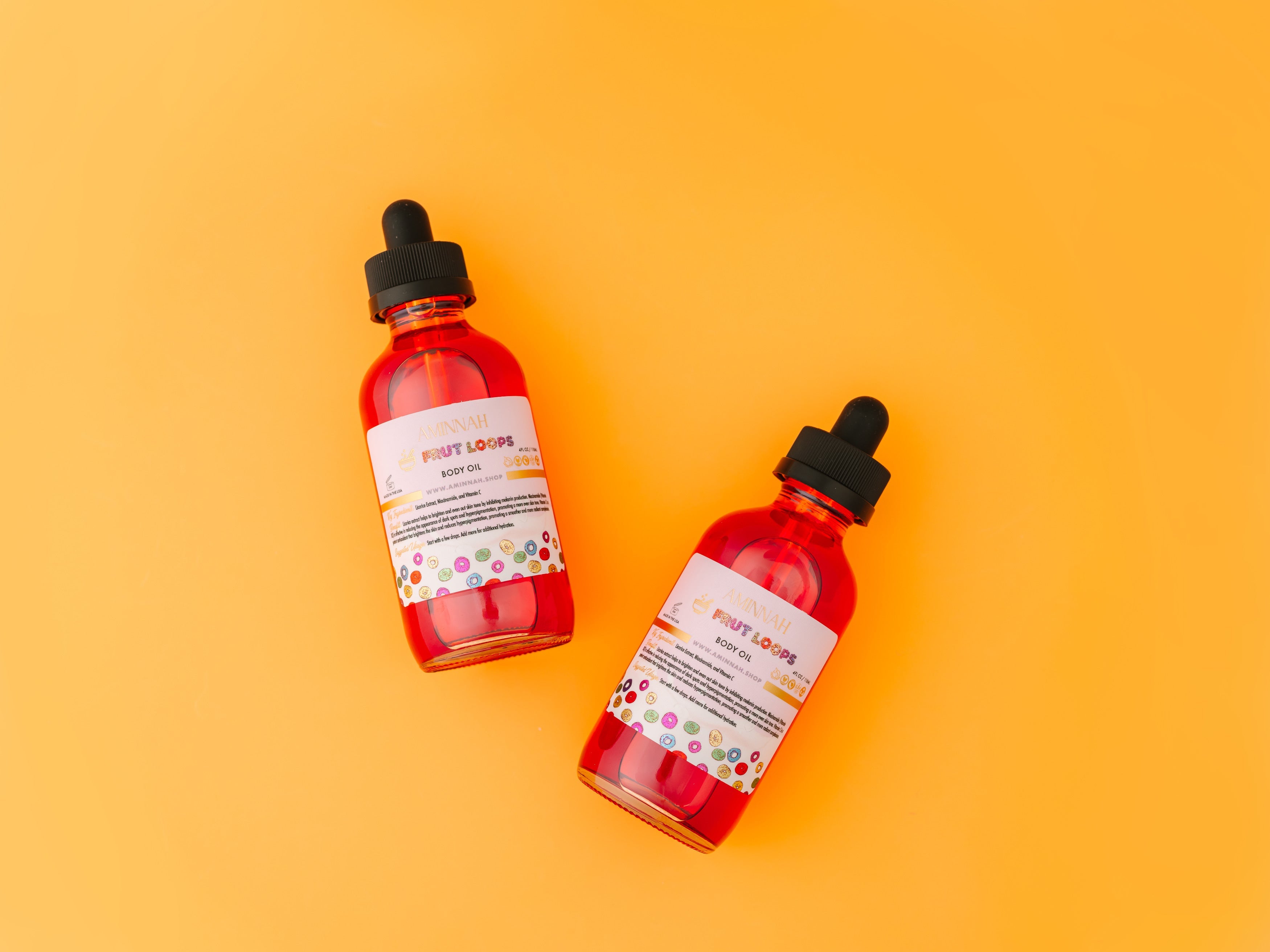 Two bottles of AMINNAH Frut Loops Body Oil with dropper applicators displayed on a bright yellow background. Clean beauty, cruelty-free, vegan body oil made with natural ingredients including Golden Jojoba Oil, Coconut Oil, Rosehip Oil, Avocado Oil, Sunflower Oil, Vitamin E, Licorice Extract, Niacinamide, and Vitamin C from a female-owned brand.