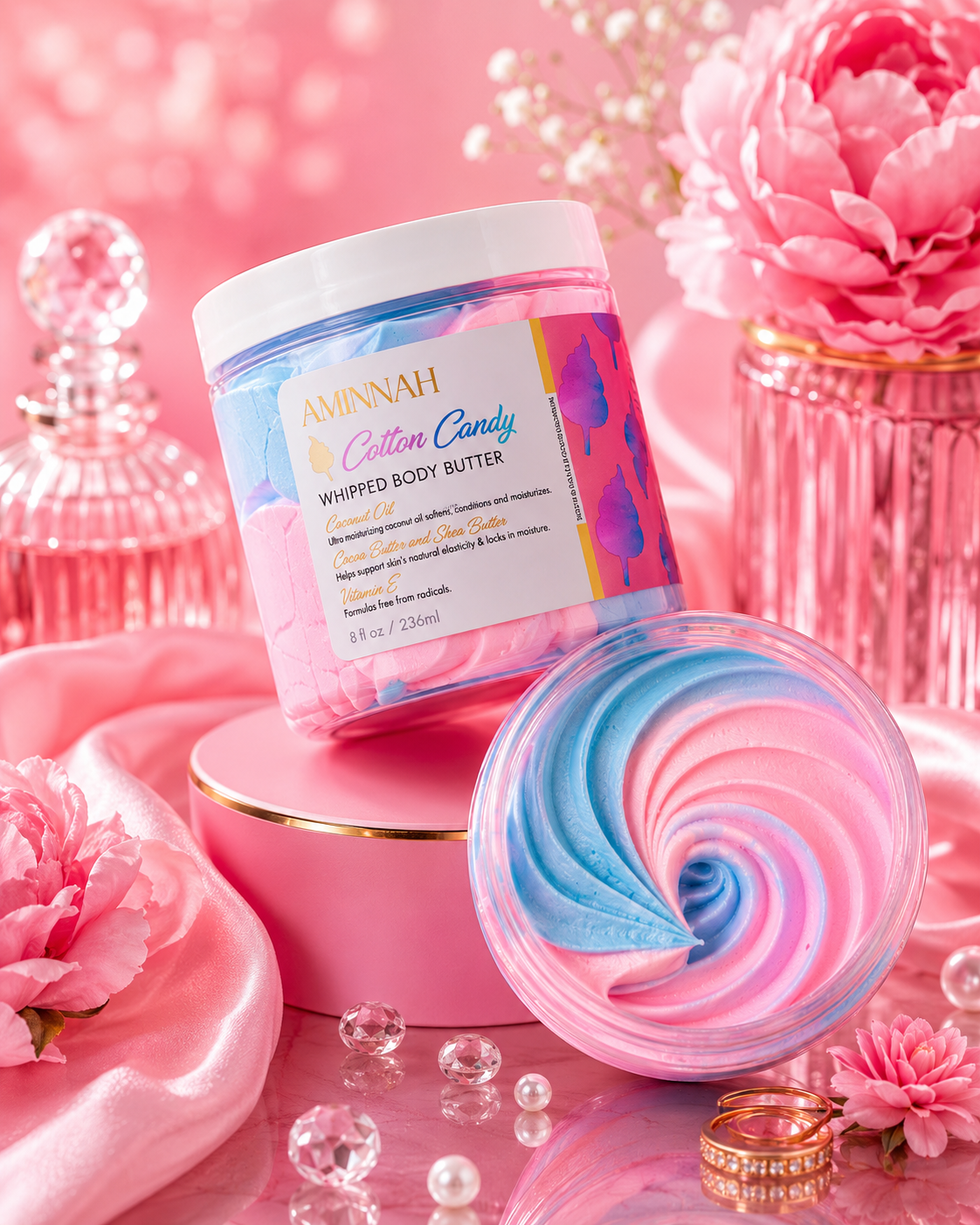 A luxurious pink display featuring the original AMINNAH Cotton Candy Whipped Body Butter in its labeled jar with a matching swirl of pink and blue whipped body butter. Styled with elegant pink flowers, crystal accents, pearls, and glossy décor to highlight a premium clean beauty skincare product from a female-owned brand. Made with natural ingredients, vegan, cruelty-free, paraben-free, and gluten-free, with recyclable packaging for eco-conscious self-care.