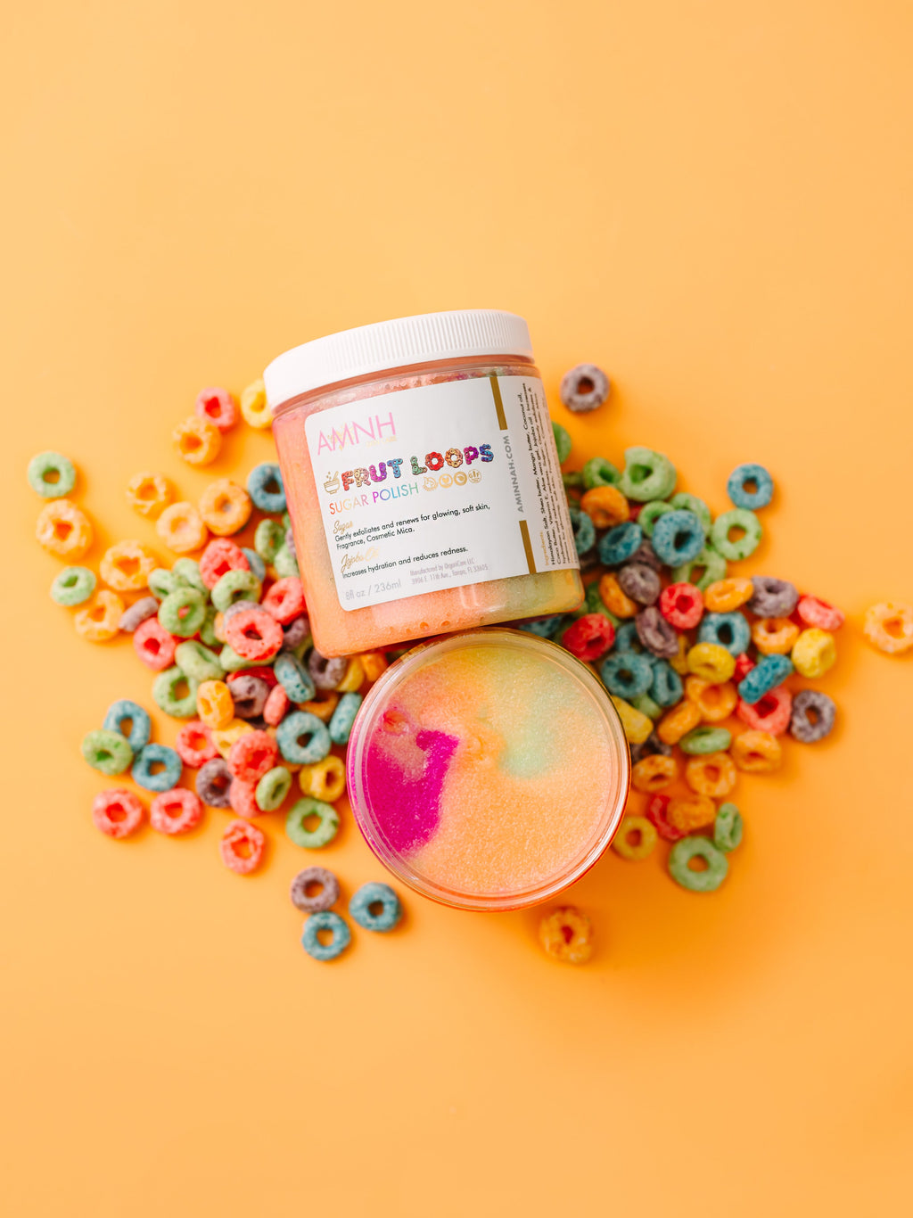 Two containers of 'Coconut Loops' bath product surrounded by colorful cereal loops on a yellow background. AMINNAH Frut Loops Sugar Polish shown open and closed on a bright yellow background surrounded by colorful cereal pieces. Clean beauty, cruelty-free, vegan sugar scrub made with naturally derived ingredients to gently exfoliate, polish away dull skin, and leave skin feeling soft, smooth, radiant, and refreshed. Female-owned brand available at Tree of Life Grocers.