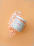 Jar of peach body scrub with a decorative label on an orange background. AMINNAH Peach Booty Tightening & Lifting Scrub in a clear jar with peach-colored exfoliating scrub displayed on a soft peach background. Clean beauty, cruelty-free, vegan body scrub made with natural ingredients including Peach Extract, Aloe Vera, Retinol, and Apricot Seed Powder from a female-owned brand.