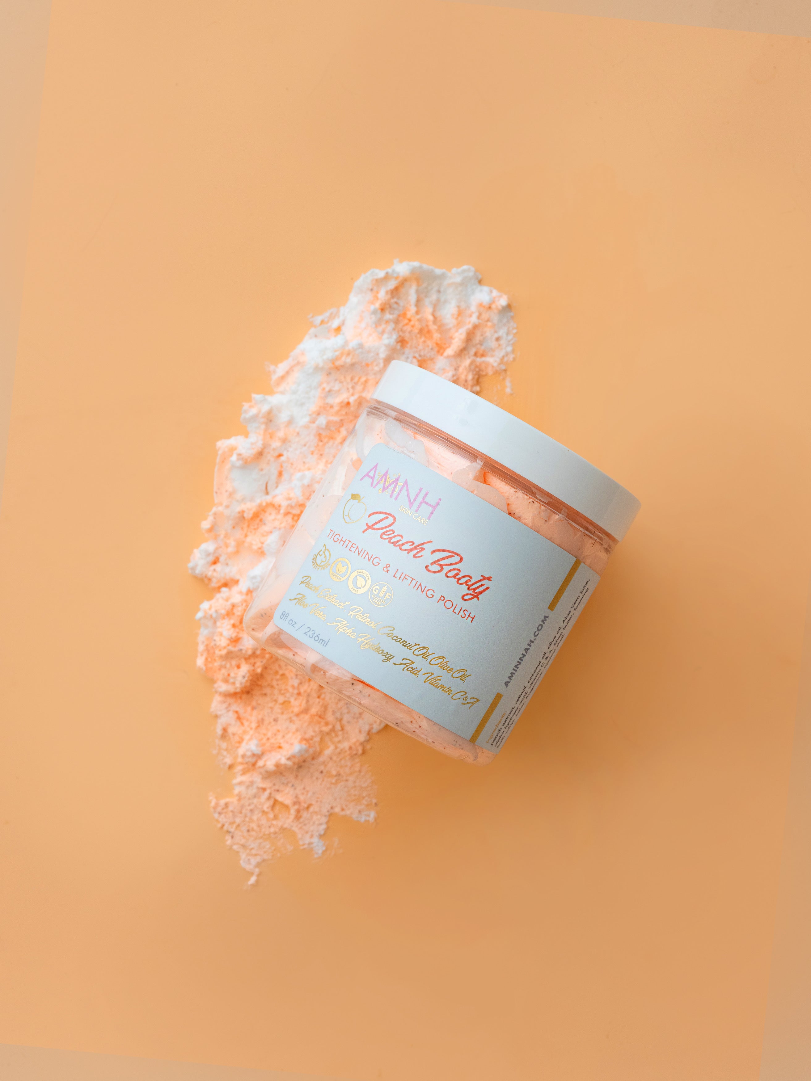 Jar of peach body scrub with a decorative label on an orange background. AMINNAH Peach Booty Tightening & Lifting Scrub in a clear jar with peach-colored exfoliating scrub displayed on a soft peach background. Clean beauty, cruelty-free, vegan body scrub made with natural ingredients including Peach Extract, Aloe Vera, Retinol, and Apricot Seed Powder from a female-owned brand.