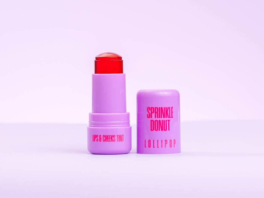 Two purple lip balm sticks with red caps on a light purple background. AMINNAH Sprinkle Donut LOLLIPOP Lips & Cheeks Tint in Hot Pink with open jelly tint stick and cap. Vegan, cruelty-free clean beauty makeup enriched with Aloe Vera, Niacinamide, Hyaluronic Acid, and Goji Berry from a female-owned brand.