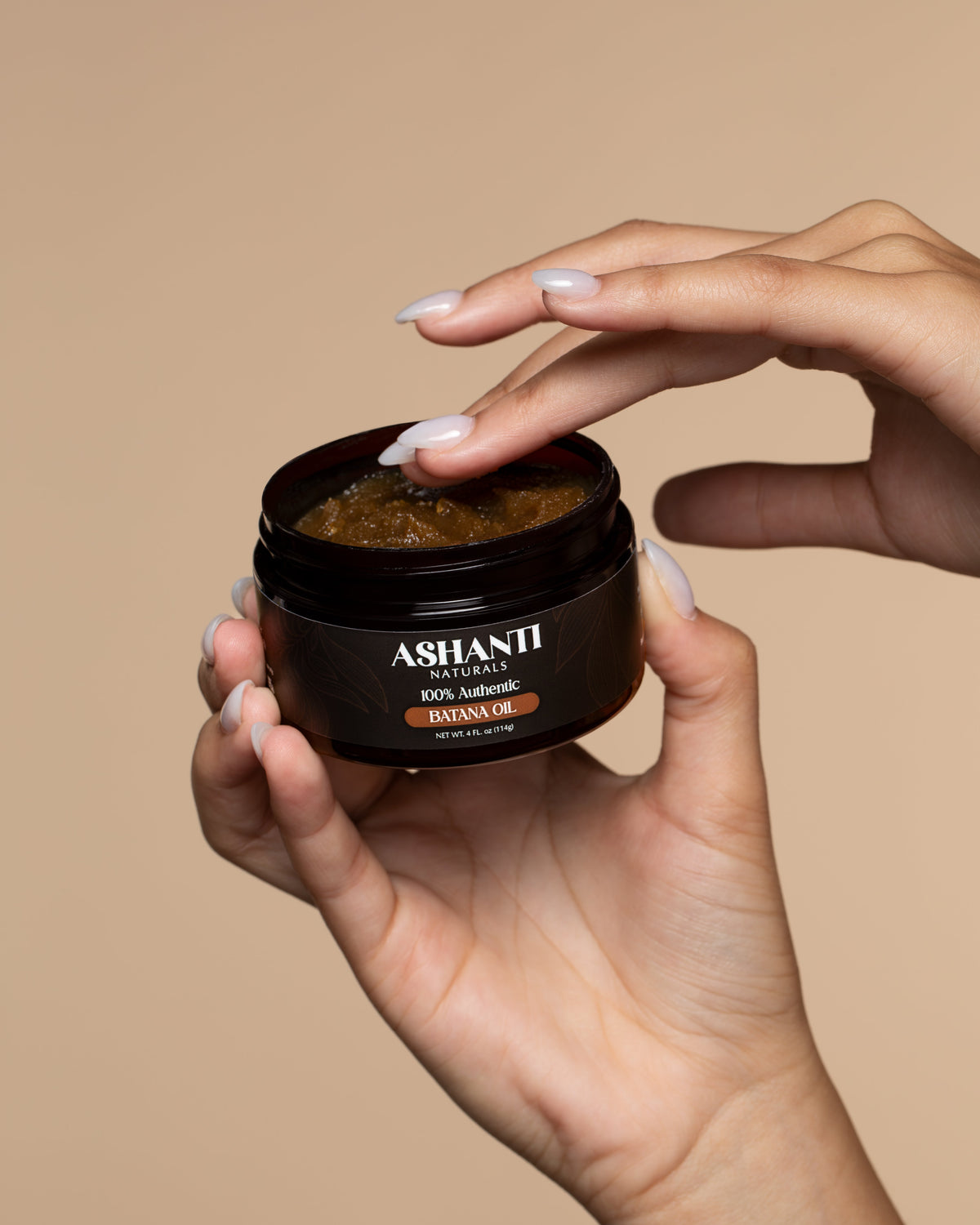 Hand holding an open jar of **Ashanti Naturals 100% Authentic Batana Oil**, showing the rich natural oil texture. Moisturizing Honduran batana oil for hair growth support, dry damaged hair repair, scalp care, strengthening, shine, and deep conditioning against a beige background.