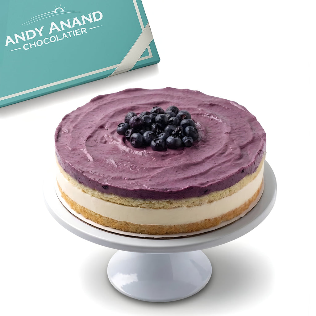 Purple frosted cake with blueberries on a white stand, Andy Anand Chocolatier branding in the background.