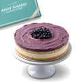 Purple frosted cake with blueberries on a white stand, Andy Anand Chocolatier branding in the background.