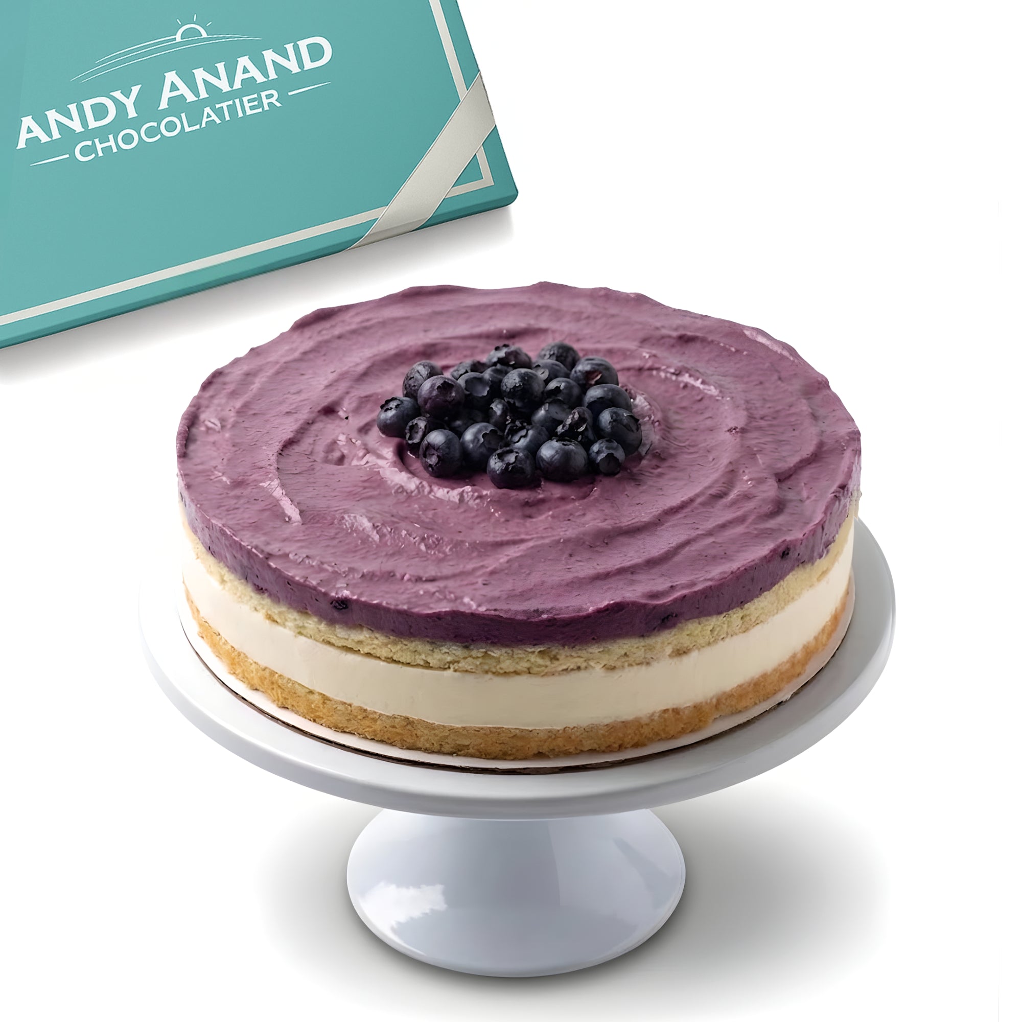 Purple frosted cake with blueberries on a white stand, Andy Anand Chocolatier branding in the background.