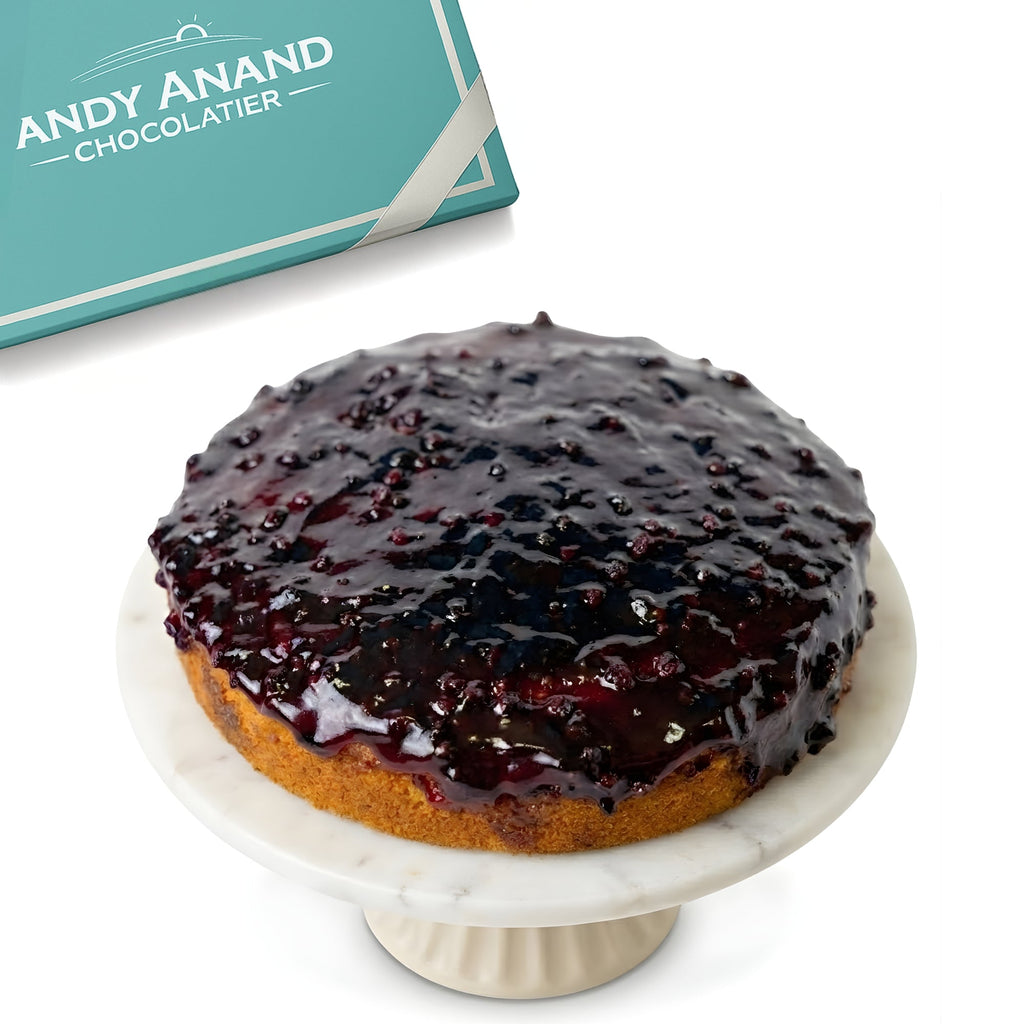 Cake with blueberry glaze on a marble stand with 'Andy Anand Chocolatier' branding in the background.