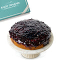 Cake with blueberry glaze on a marble stand with 'Andy Anand Chocolatier' branding in the background.