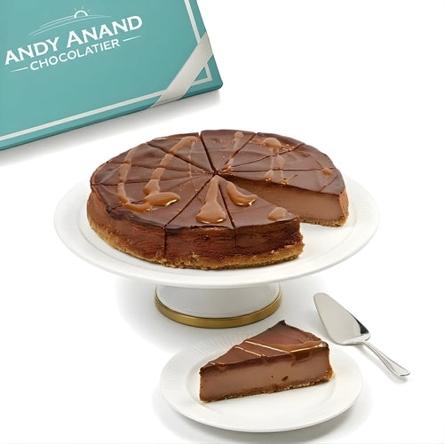 Chocolate cake with a slice taken out, displayed on a white cake stand with a 'Andy Anand Chocolatier' box in the background.