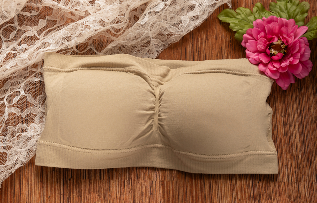 Beige seamless strapless bandeau bra with gathered center detail, soft stretch fabric, delicate white lace and pink flower on rustic wood background, women’s lingerie, neutral nude bra, comfortable wireless bralette, feminine intimate apparel, romantic floral styling, elegant everyday undergarment product photography, Tree of Life Grocers lingerie and self-care collection