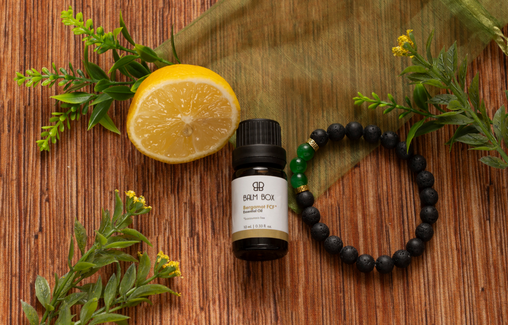 Balm Box Bergamot Essential Oil bottle with natural lemon, fresh green botanical leaves, and black and green gemstone bracelet on rustic wooden background, aromatherapy essential oil and natural wellness self-care product for relaxation, mindfulness, holistic living, home spa, beauty and wellness gifts from Tree of Life Grocers