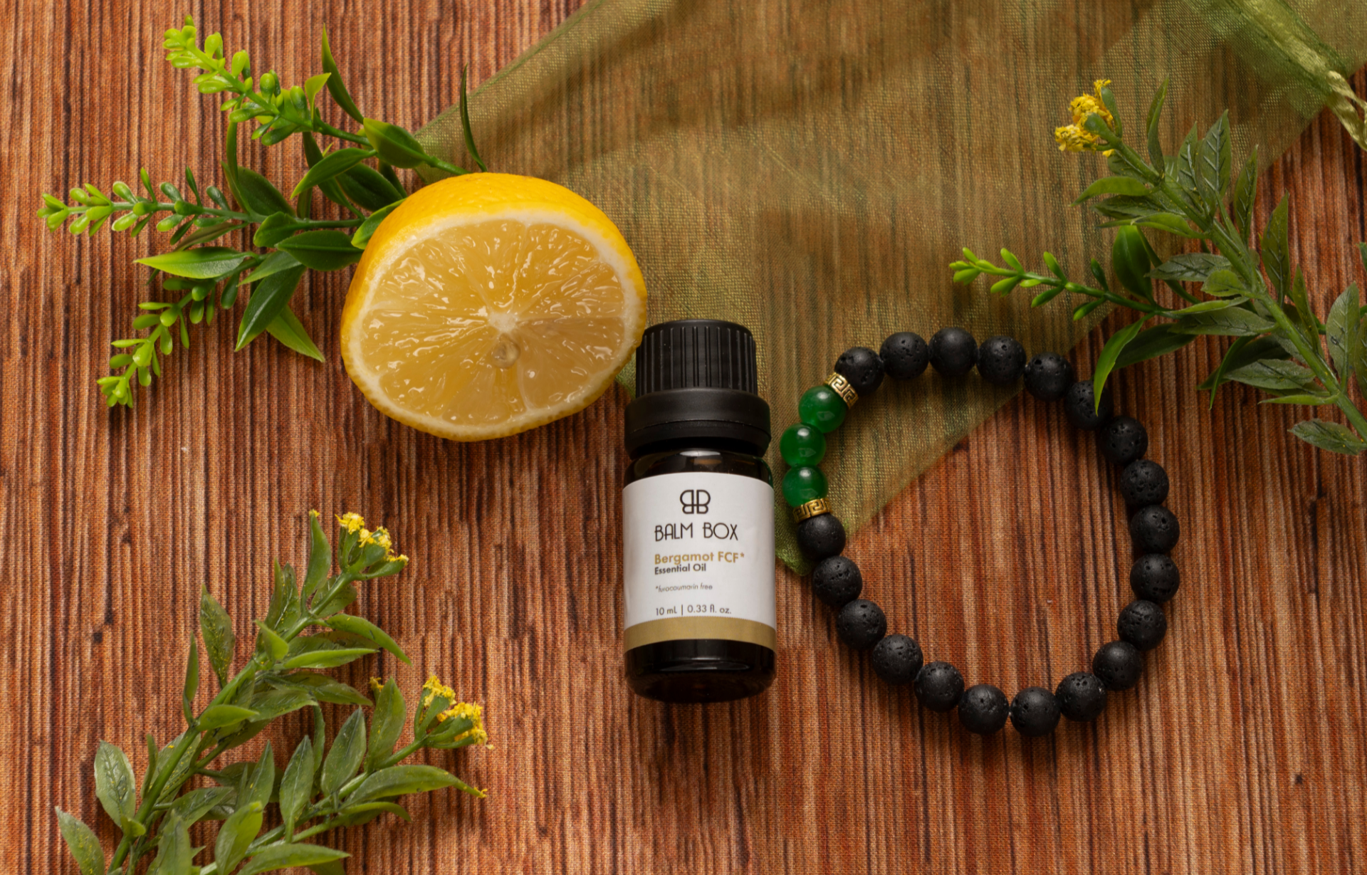 Bottle of Bergamot Essential Oil with green leaves, fresh lemon, black and green beaded bracelet, natural aromatherapy and self-care gift set on rustic wood background.