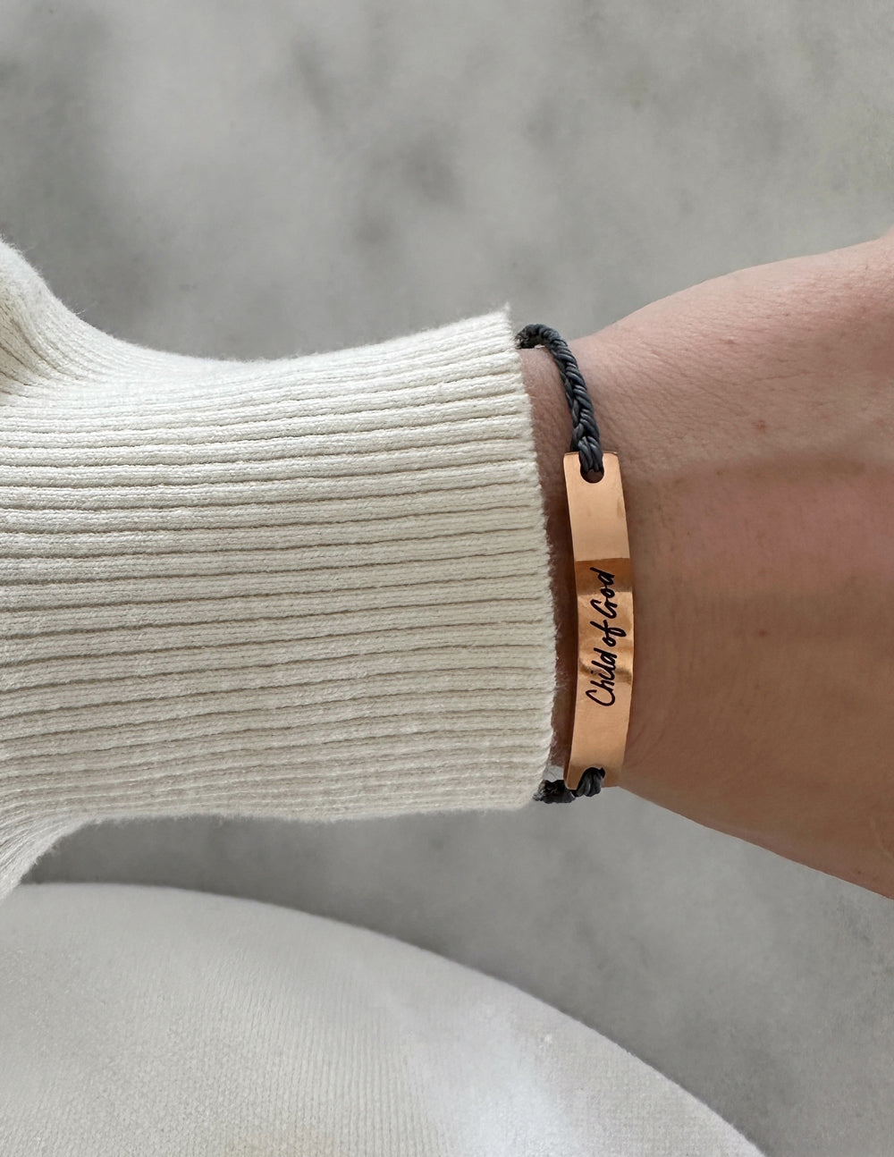 A close-up shot of a rose gold bracelet featuring engraved text, worn on a wrist against a clean, neutral background. Chid of God Bracelet.