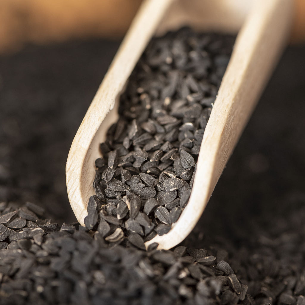 Organic black cumin (Nigella sativa) seeds in a wooden scoop on a bed of black cumin seeds.
