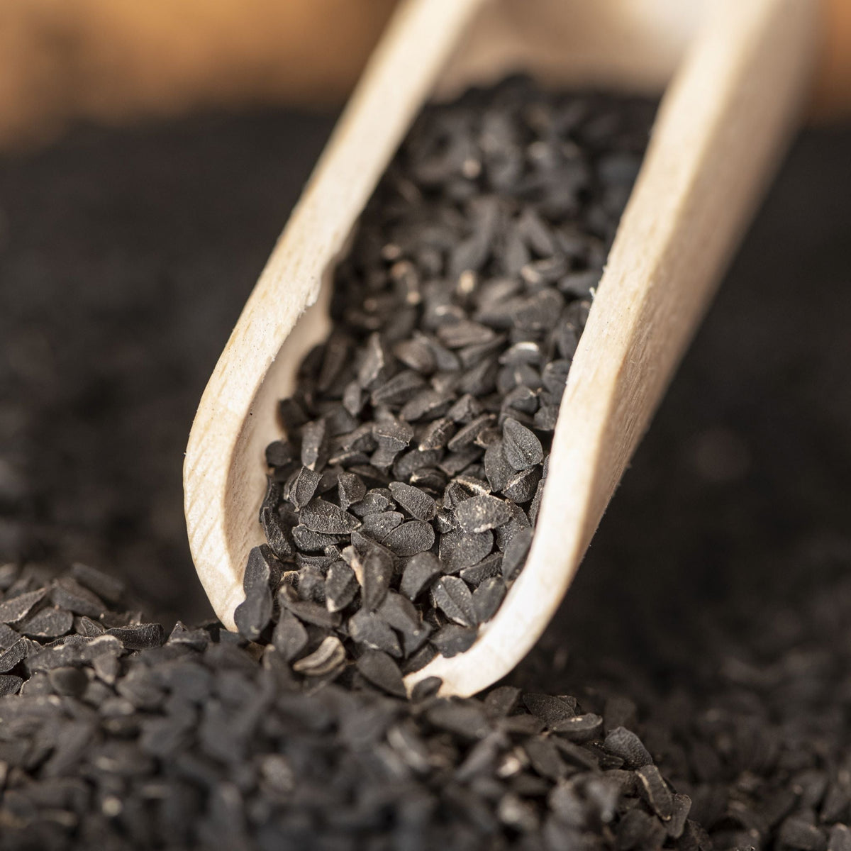 Organic black cumin (Nigella sativa) seeds in a wooden scoop on a bed of black cumin seeds.