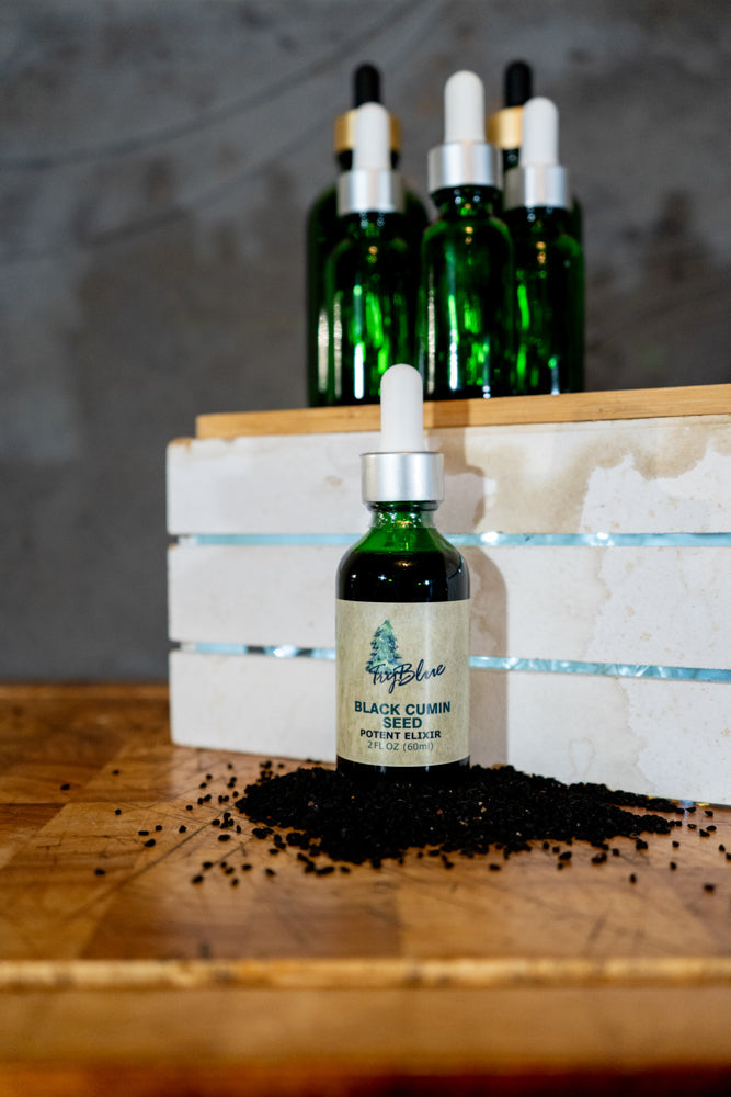 Black Cumin Seed Elixir dropper bottle displayed with black cumin seeds on a wooden surface, with additional green glass dropper bottles in the background.