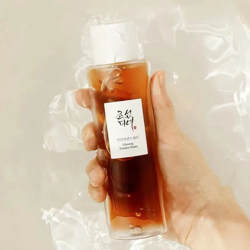 Hand holding a bottle of **Beauty of Joseon Ginseng Essence Water** with amber essence, white cap, and Korean label against a soft, light background with shimmering water reflections.