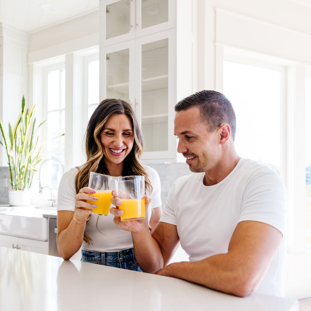 Man and woman holding glasses of orange juice in a bright kitchen.