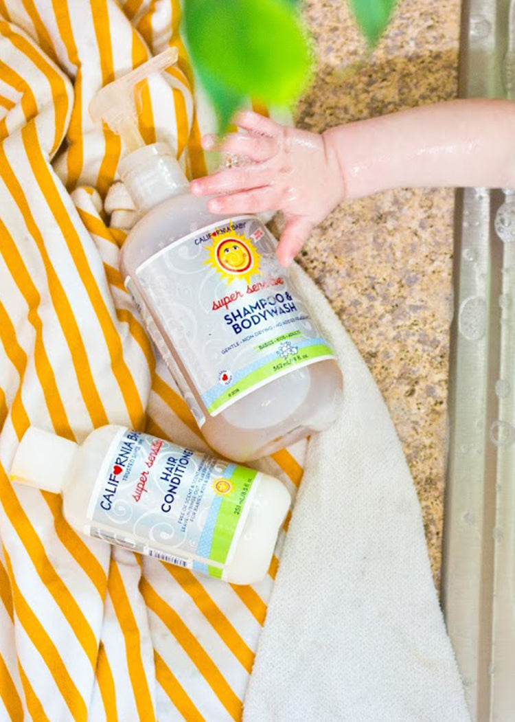 Child's hand holding a bottle of shampoo and body wash with a yellow and white striped towel in the background. California Baby® (No Fragrance) Super Sensitive™ Hair Conditioner - USDA Certified Biobased Product 100% Plant-Based