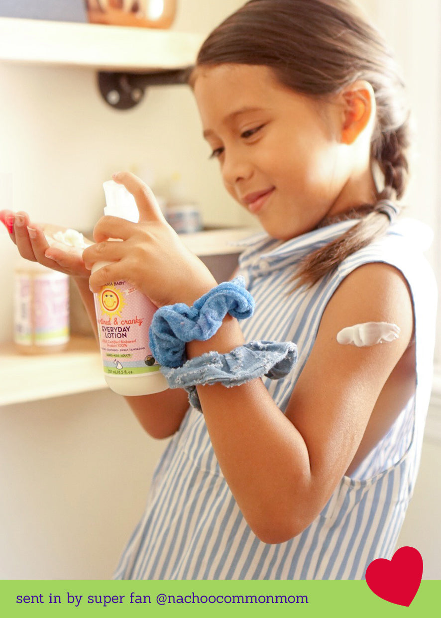 Young girl smiling as she applies lotion to her arm while holding a bottle of California Baby Overtired & Cranky™ Everyday Lotion for Sensitive Skin, 8.5 oz (251 mL), in a bright kitchen setting.