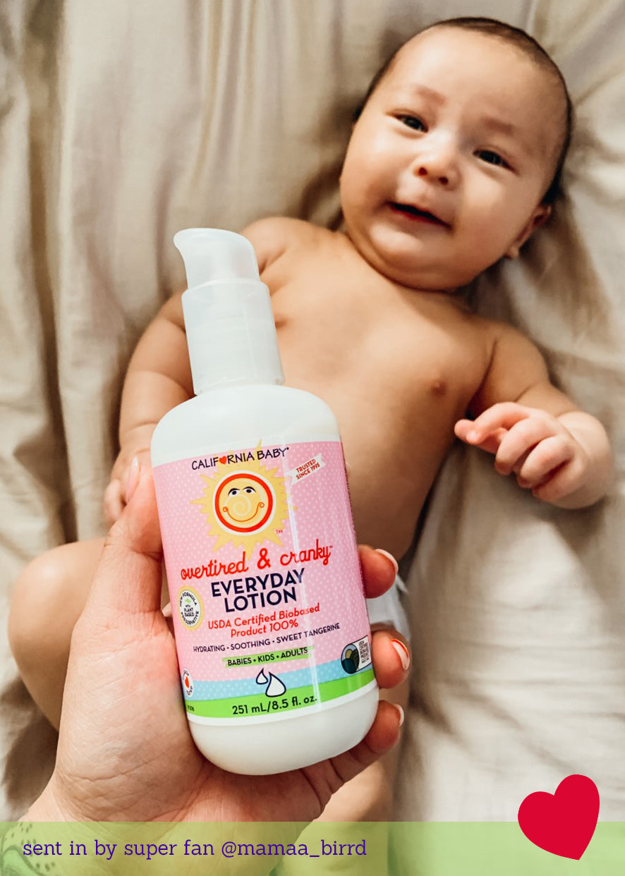 Baby holding a bottle of California Baby Overtired & Cranky™ Everyday Lotion for Sensitive Skin (8.5 oz) while lying on a bed, with a red heart graphic in the bottom-right corner.